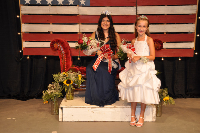 girl with crown and bouquet of flowers sitting in front of american flag with younger girl