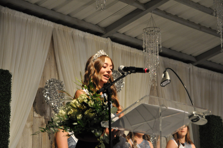 girl with crown and bouquet of flowers speaking at podium